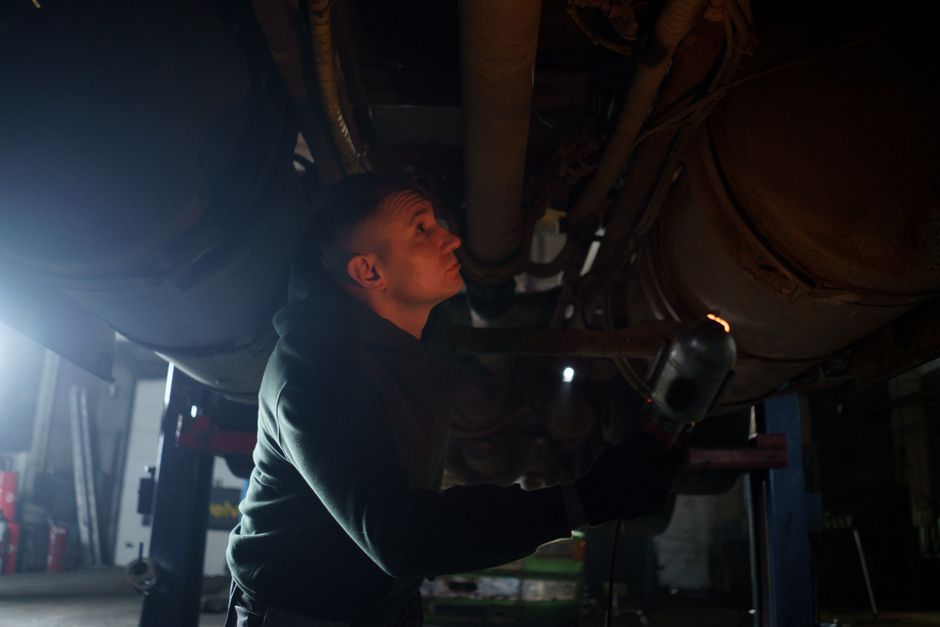 Auto mechanic inspects vehicle underbody in a garage setting with dim lighting.